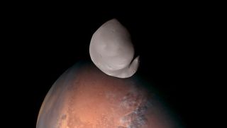 The smooth, gray moon Deimos hovers in front of the rocky red surface of Mars