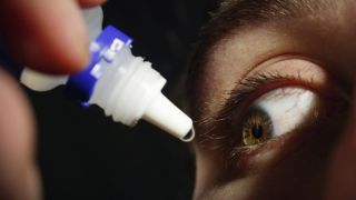 close up of a man's eye as he's squeezing a generic eye drop dropper near his face, pushing out a drop of clear liquid