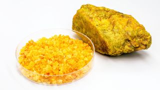 An Uranium nitrate called uranyl in a bowl; looks like a bowl of yellow aquarium pebbles. A solid chunk of bright yellow uranium ore sits to the right of the bowl on a white background